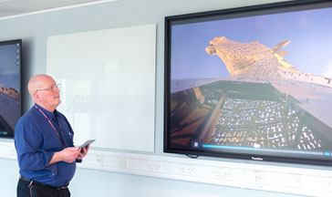 A ֱ staff member standing by 2 screens showing the Scottish Kelpies sculpture