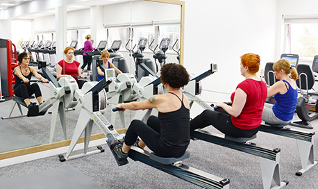 A row of women using rowing machines in front of a mirrored wall, ֱ