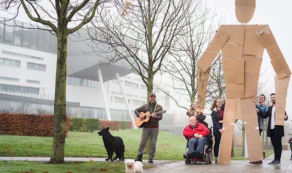 Students outside ֱ holding a giant card board person up whilst a man with a dog plays guitar