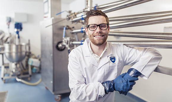 ֱ student smiling at the camera in a lab coat and protective gloves